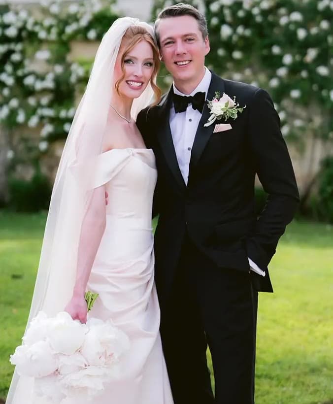 The bride in a strapless gown and long veil, holding white peonies, beside the groom in a black tuxedo with a pink boutonniere, on a lawn before a wall of white roses