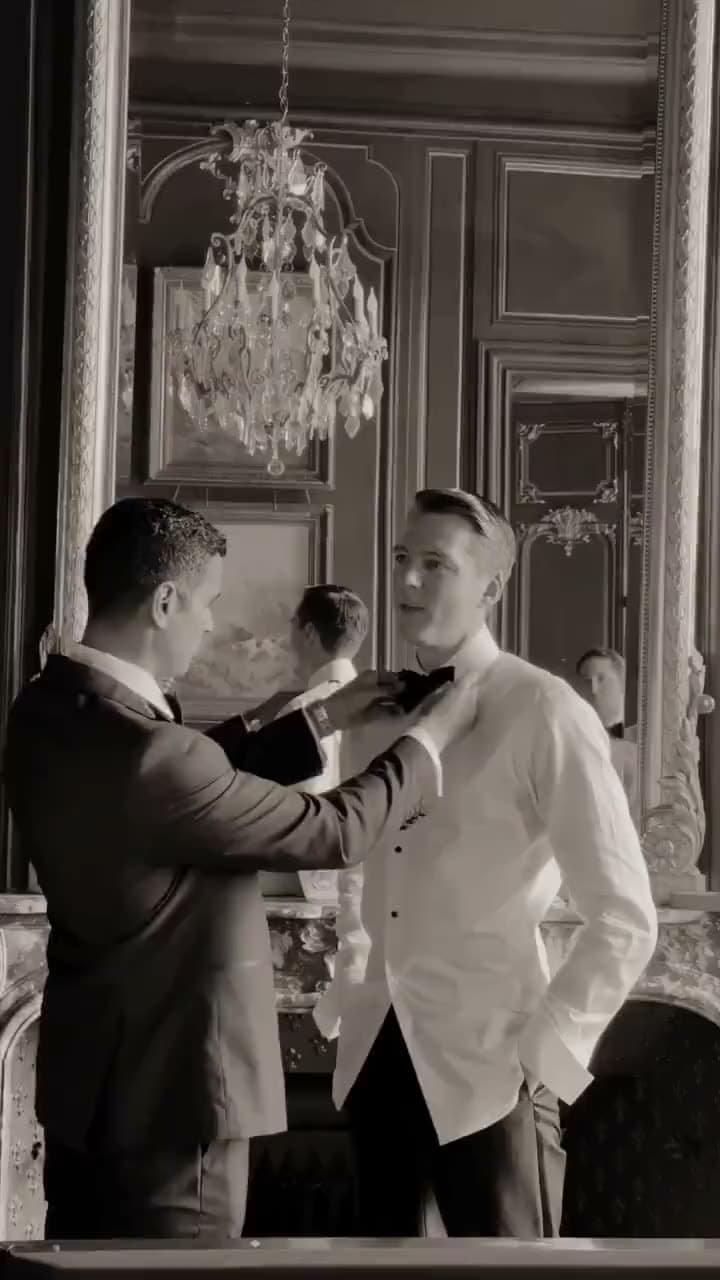 A groomsman tying the groom's black bow tie in a gilded mirror beneath a crystal chandelier