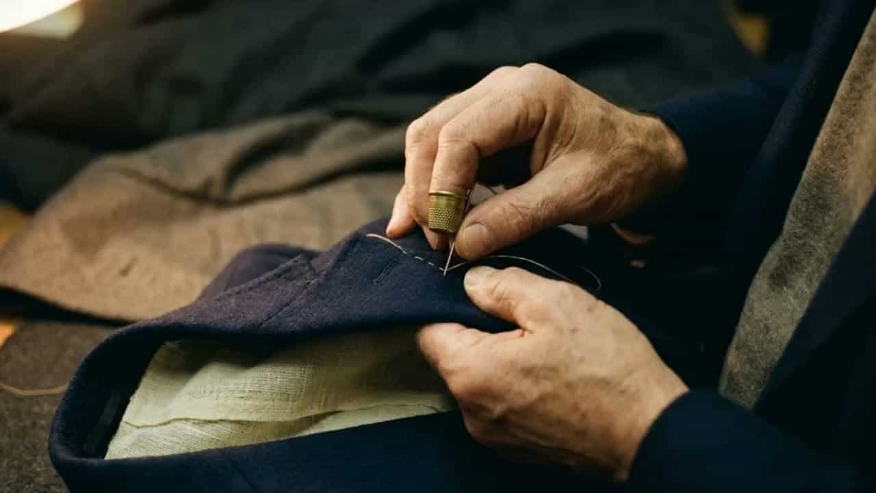 Close-ups from a tailor's workroom: hands hand-stitching navy cloth, bolts of wool, a buttonhole, swatch books, shears and chalk, and a basted jacket on a form