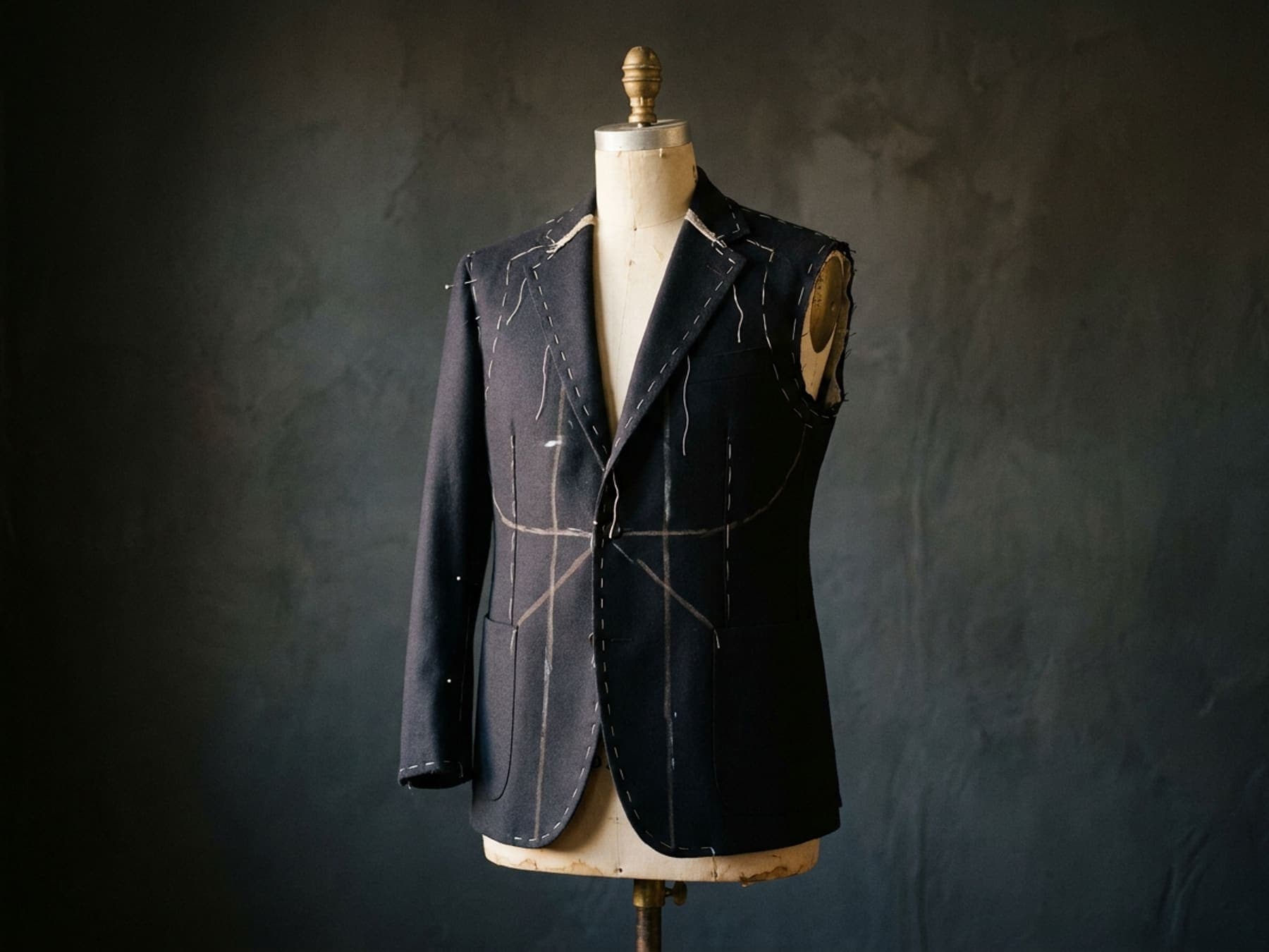 A navy jacket on a dress form, held together with white basting thread and chalk lines